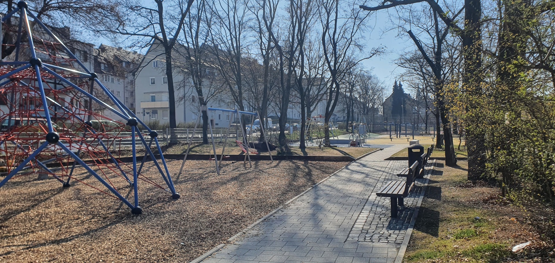 Blick auf den Spielplatz: Links sind die Spielgeräte zu sehen. In der Mitte des Bildes ist ein gepflasterter Weg  an dem rechts Sitzbänke aufgestellt sind. Der Spielplatz ist links und rechts von Bäumen gesäumt.