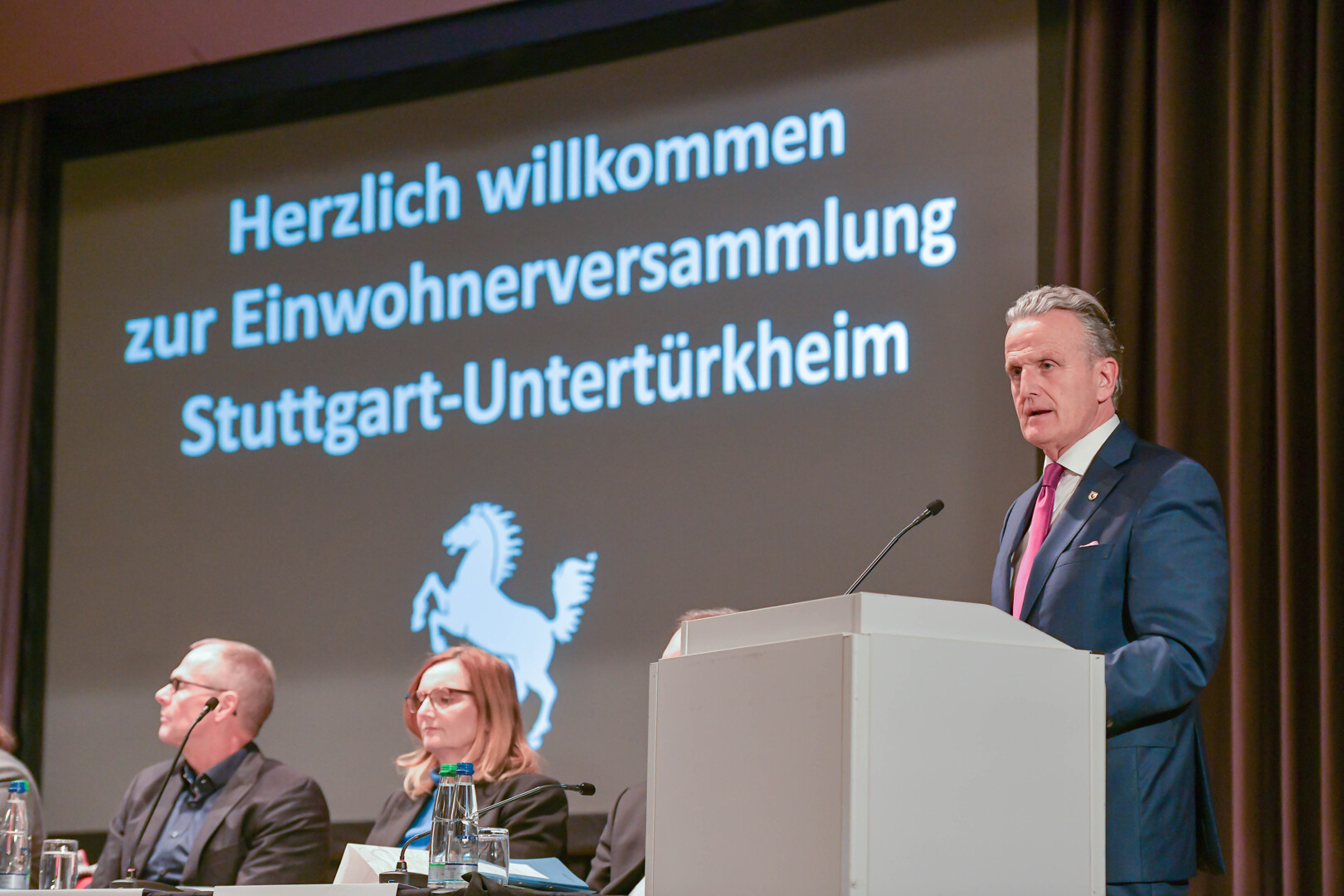 Ein Mann steht am Rednerpult. Auf einer Leinwand im Hintergrund steht "Herzlich willkommen zur Einwohnerversammlung Stuttgart-Untertürkheim"